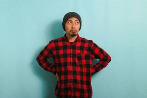 A bored young Asian man wearing a beanie hat and a red plaid flannel shirt stands with an arm akimbo gesture, looking to the right deep in thought about what to do, isolated on a blue background photo