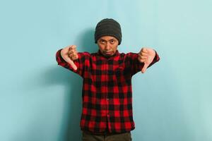 Annoyed Young Asian man with beanie hat and red plaid flannel shirt showing a thumbs-down gesture, giving a bad review, looking disappointed, frowning, and feeling sad, isolated on blue background photo