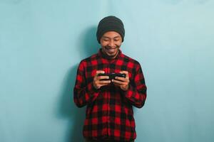 Excited young Asian man with a beanie hat and a red plaid flannel shirt is playing an online game on his smartphone, isolated on a blue background photo