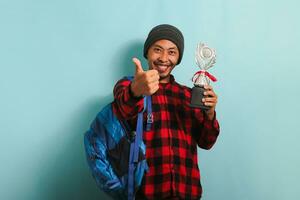 Excited Young Asian man student wearing backpack, beanie hat, and red plaid flannel shirt, giving thumbs-up while holding trophy, rejoicing in his success and achievement, isolated on blue background photo