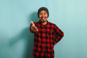 A young Asian man with a beanie hat and a red plaid flannel shirt is ready to shake hands while standing against a blue background photo