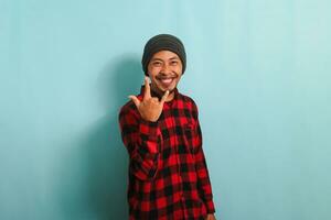 Happy young Asian man with a beanie hat and red plaid flannel shirt showing the rock and roll gesture with his fingers, expressing love for heavy metal music, isolated on a blue background photo