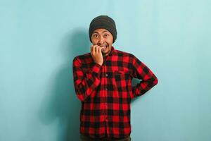 Young Asian man with a beanie hat and red plaid flannel shirt bites his fingernails in puzzlement, looking nervously at the camera, isolated on a blue background photo