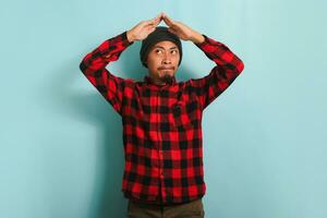 Pensive young millennial Asian man with a beanie hat and red plaid flannel shirt making a roof gesture above his head, thinking and planning to have his own home, isolated on a blue background. photo