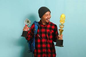 Happy young Asian man student wearing a backpack, beanie hat, and red plaid flannel shirt, lifting up his trophies, rejoicing in his success and achievement, isolated on a blue background. photo