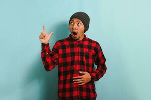 Surprised Young Asian man with a beanie hat and red plaid flannel shirt pointing his finger aside at empty copy space, isolated on a blue background photo