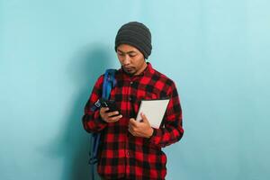 Pensive young Asian student with a beanie hat and a red plaid flannel shirt, wearing a backpack, is searching for ideas on the internet to finish his assignment isolated on blue background photo