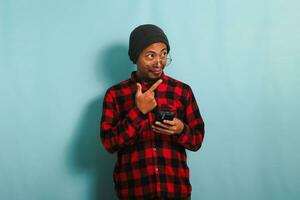 Happy young Asian man wearing eyeglasses, a beanie hat, and a red plaid flannel shirt is pointing his finger aside at copy space while holding a smartphone, isolated on a blue background photo