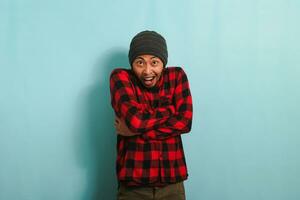 Excited Young Asian man with beanie hat and red plaid flannel shirt holds his hands crossed, his mouth wide open in surprise, isolated on a blue background photo