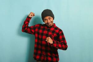 Confident Young Asian man with beanie hat and red plaid flannel shirt making a strong gesture, showing strength by flexing his arms and muscles, isolated on a blue background photo