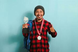 Excited young Asian man student wearing backpack, beanie hat, and red plaid flannel shirt, giving thumbs up while holding a trophy and rejoicing in success and achievement, isolated on blue background photo