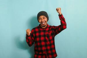 Enthusiastic young Asian man with beanie hat and red plaid flannel shirt rejoicing, saying yes, looking happy, and celebrating victory with a champion fist pump gesture, isolated on blue background photo