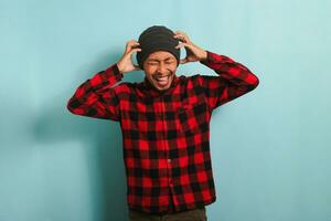 Excited young Asian man with beanie hat and red plaid flannel shirt grabs his head, stands in surprise and speechlessness, receiving incredible news while standing against a blue background photo