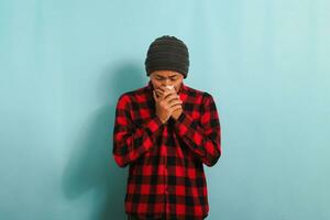 A crying young Asian man with a beanie hat and a red plaid flannel shirt wipes tears with a tissue while standing against a blue background photo