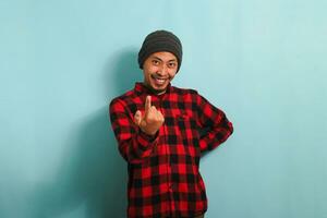 A young Asian man wearing a beanie hat and a red plaid flannel shirt is making a Come Here gesture with his hand, inviting, welcoming, happy, and smiling. He is isolated on a blue background photo