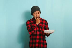 Pensive Young Asian man with beanie hat and red plaid flannel shirt is feeling confused, looking aside, thinking about what to eat while holding an empty white plate, isolated on a blue background photo
