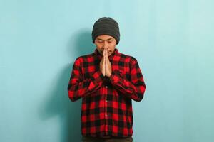 A young Muslim Asian man with a beanie hat and red plaid flannel shirt is praying to Allah, the Muslim God, keep eyes closed, isolated on a blue background photo