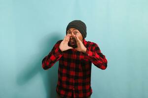 A young Asian man with a beanie hat and a red plaid flannel shirt keeps his hand on his mouth, telling a secret and spreading a rumor through whispering while standing against a blue background photo