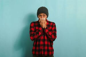 A young Asian man wearing a beanie hat and a red plaid flannel shirt is sneezing into a tissue, suffering from a cough, fever, and flu. He is isolated on a blue background photo