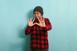 Ew, It's so gross. An annoyed young Asian man with a beanie hat and a red plaid flannel shirt is repulsed by something, displaying a disgusted expression while standing against a blue background photo