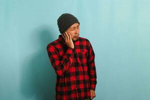 Young Asian man with beanie hat and red plaid flannel shirt holds his hand near his ear, suffering from tinnitus, a throbbing earache, and fatigue due to noise, while standing against blue background photo