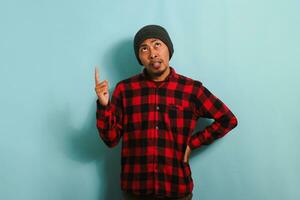 A confused young Asian man with a beanie hat and a red plaid flannel shirt is pointing and looking up at the copy space with a frowning and bothered expression while standing against a blue background photo