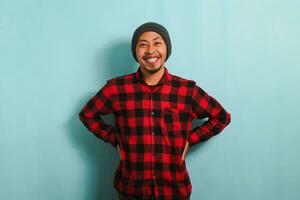 A confident young Asian man with a beanie hat and a red plaid flannel shirt is keeping his hand on his waist in an akimbo gesture. He is isolated on a blue background photo