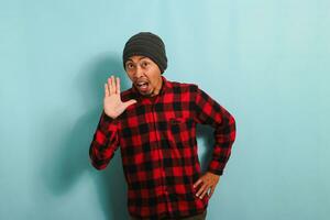 Excited Young Asian man with a beanie hat and red plaid flannel shirt shouting and holding his palm near his opened mouth, telling good news, isolated on a blue background photo