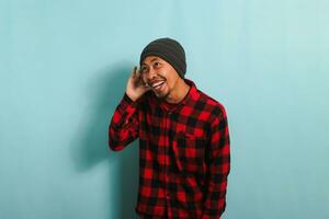 A young Asian man with a beanie hat and a red plaid flannel shirt smiles while placing his hand over his ear, listening to a rumor or gossip, while standing against a blue background photo