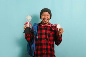 Happy young Asian man student wearing a backpack, beanie hat, and red plaid flannel shirt, showing a medal and trophy while rejoicing in success and achievement, isolated on a blue background photo