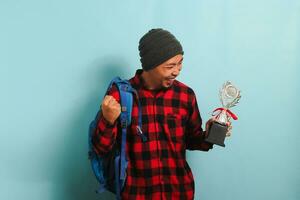 Excited young Asian man student wearing a backpack, beanie hat, and red plaid flannel shirt, making a YES gesture while holding a trophy and rejoicing in achievement, isolated on a blue background photo