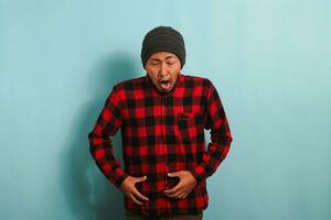 An annoyed young Asian man with a beanie hat and a red plaid flannel shirt feels nauseous as he smells an unpleasant and disgusting odor while standing against a blue background photo