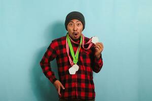 Excited young Asian man with a beanie hat and a red plaid flannel shirt is amazed, with his mouth open in surprise and disbelief, as he wins medals while standing against a blue background photo