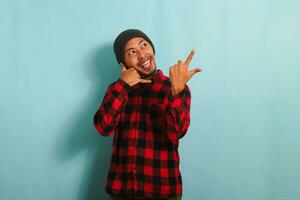 un emocionado joven asiático hombre vistiendo un gorro sombrero y un rojo tartán franela camisa hace un llamada yo gesto, señalando a el Derecha espacio de copia, aislado en un azul antecedentes foto