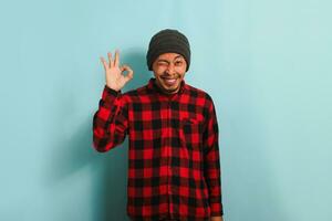 Smiling, winking young Asian man with beanie hat and red plaid flannel shirt does the OK sign gesture in approval as he says good job or well done, expressing satisfaction, isolated on blue background photo