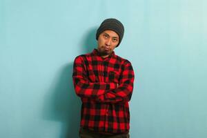 An angry young Asian student man wearing a beanie hat and a red plaid flannel shirt frowns and keeps his arms folded, appearing bored, isolated on a blue background photo