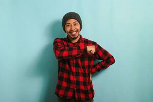 A confident young Asian man with a beanie hat and a red plaid flannel shirt keeps his fist on his chest, feeling proud, while standing against a blue background photo