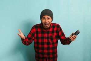 Excited young Asian man with a beanie hat and red plaid flannel shirt watching TV and cheering for a sports competition with laughter and a sense of victory, isolated on a blue background photo