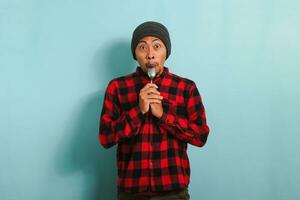 Surprised Young Asian man with beanie hat and red plaid flannel shirt holding a spoon near his mouth, feeling excited and eager to eat, isolated on a blue background photo