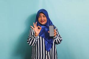 An excited middle-aged Asian woman in a blue hijab and a striped shirt is holding her phone with a blank white screen while making an approval sign or OK gesture. She is isolated on a blue background. photo