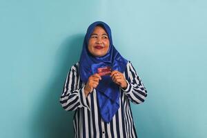 Excited Middle-aged Asian woman in blue hijab and striped shirt showing, presenting credit card for paying online business, isolated over blue background. photo