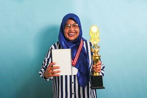 A happy middle-aged Asian businesswoman in a blue hijab and a striped shirt is holding an empty white book and gold trophy, celebrating her success, isolated on a blue background photo
