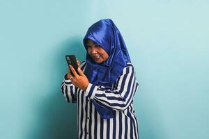 An angry middle-aged Asian woman in a blue hijab and a striped shirt is reacting to bad news on her phone, expressing shock and anger with her gestures. She is isolated on a blue background photo