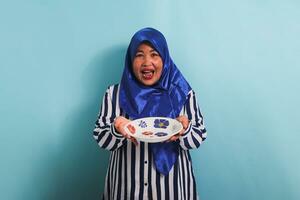 An excited middle-aged Asian woman in a blue hijab and a striped shirt is standing and holding an empty white plate. She is isolated on a blue background photo