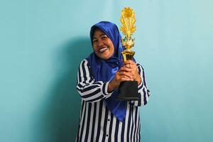 un emocionado de edad mediana asiático mujer de negocios en un azul hijab y un a rayas camisa es participación un oro trofeo, celebrando su éxito y logro. ella es aislado en un azul antecedentes foto