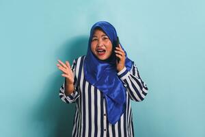 A shocked middle-aged Asian woman in a blue hijab and a striped shirt is talking on the phone with a surprised expression, hearing big news. She is isolated on a blue background photo