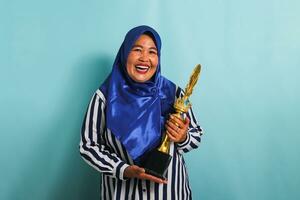 un emocionado de edad mediana asiático mujer de negocios en un azul hijab y un a rayas camisa es sonriente mientras participación un oro trofeo, celebrando su éxito y logro. ella es aislado en un azul antecedentes foto
