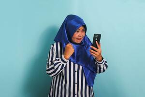 An angry middle-aged Asian woman in a blue hijab and a striped shirt is reacting to bad news on her phone, expressing shock and anger with her gestures. She is isolated on a blue background photo