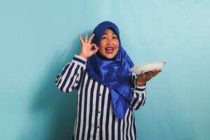 A middle-aged Asian woman in a blue hijab and a striped shirt is showing the OK sign while holding an empty white plate or dish. She is isolated on a blue background photo