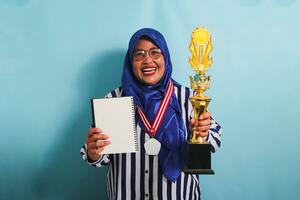 un contento de edad mediana asiático mujer de negocios en un azul hijab y un a rayas camisa es participación un vacío blanco libro y oro trofeo, celebrando su éxito, aislado en un azul antecedentes foto